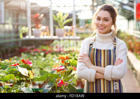 Happy charming female gardener in uniform standing near anthuriums in greenhouse Stock Photo
