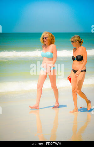 Two older middle aged women in bikinis stroll the beach conversing and enjoying the white sand and the nearby gentle waves Stock Photo