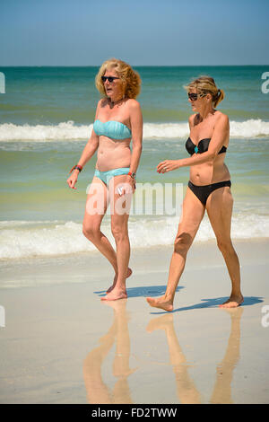 Two older middle aged women in bikinis stroll the beach conversing and enjoying the white sand and the nearby gentle waves Stock Photo