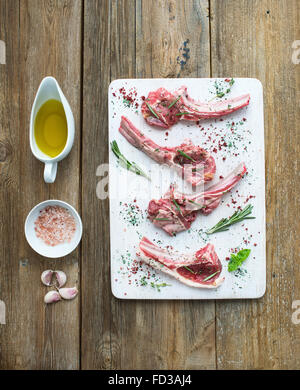 Raw lamb chops. Rack of Lamb with garlic, rosemary and spices on white chopping board, oil in a saucer, salt, dinnerware over ru Stock Photo