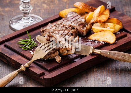Sliced well done grilled New York steak with roasted potato wedges on cutting board on wooden background Stock Photo