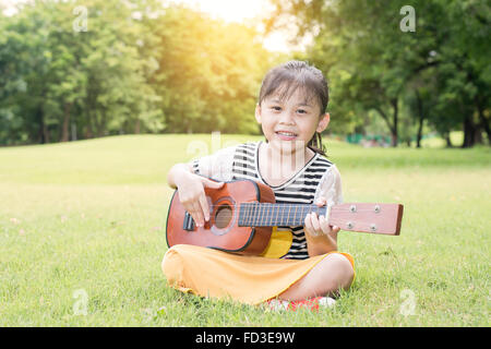 Asian little girl sitting on grass and play ukulele in park Stock Photo