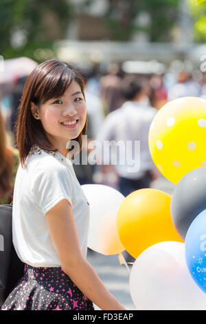 Thai girl in casual dress is smiling and holding colorful balloons in outdoor crowded place Stock Photo