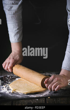Female hands rolling out dough for pasta by wooden rolling pin over wooden kitchen table, powdering by flour. Dark rustic style. Stock Photo