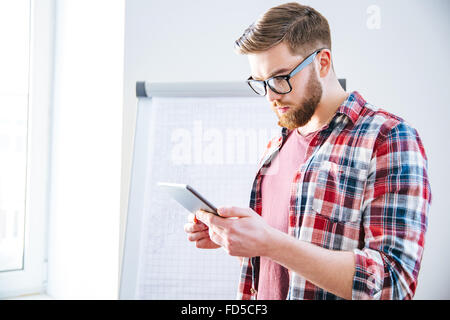 Handsome serious young man with beard in plaid shirt standing near flipchart and using tablet Stock Photo
