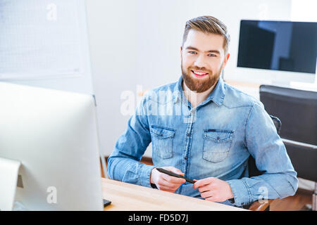Cheerful handsome young designer with beard in blue shirt holding stylus and sitting at the table in office Stock Photo