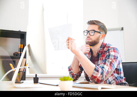 Serious focused young bearded designer in checkered shirt looking and checking blueprint in office Stock Photo