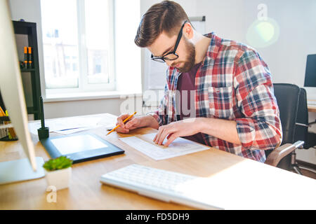 Portrait of young male designer in glasses sitting in office and making blueprint Stock Photo