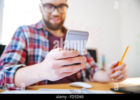 Happy attractive bearded young man in glasses sitting and using smartphone in office Stock Photo