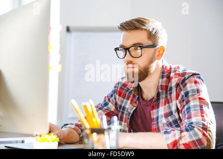 Serious handsome bearded man in checkered shirt sitting on workplace and working with computer Stock Photo