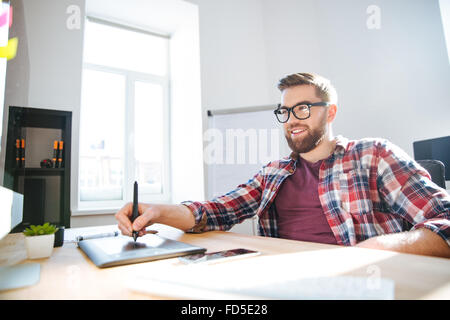Smiling handsome young bearded designer in checkered shirt and glasses drawing and using graphic pen tablet and stylus Stock Photo