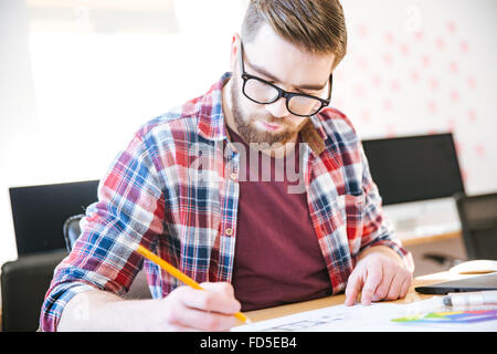 Concentrated young man with beard in plaid shirt and glasses making sketches with pencil sitting at the table Stock Photo