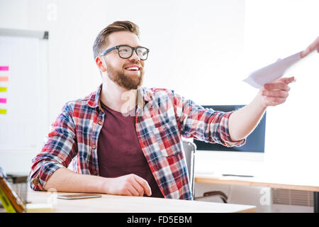 Cheerful attractive young man in checkered shirt siting on workplace and receiving documents Stock Photo