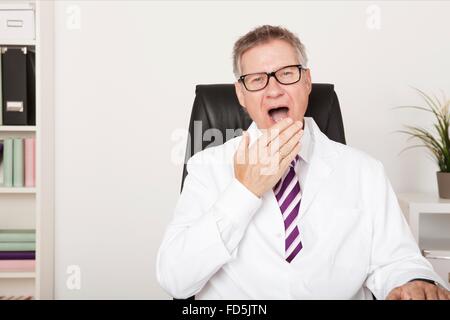 Yawning middle-aged male doctor sitting at his desk in his office tired out after a long days work and ready to head for home Stock Photo