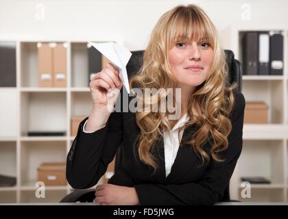 Playful attractive young blond businesswoman with a paper plane in her hand, sitting at her desk ready to launch it Stock Photo