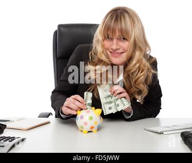 Smiling happy young businesswoman saving part of her hard earned wealth, inserting a dollar bill into a polka dot piggy bank Stock Photo