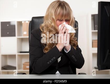 Young businesswoman blowing her nose as she sits at her desk at work suffering from seasonal hayfever, a cold or influenza Stock Photo