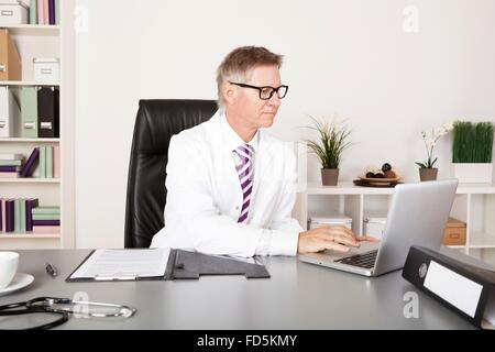 Male Doctor Using Laptop for Medical Reports Sitting on a Desk Stock Photo