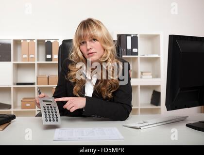 Attractive young manageress or businesswoman sitting at her desk in the office pointing to a calculator turned to the camera Stock Photo
