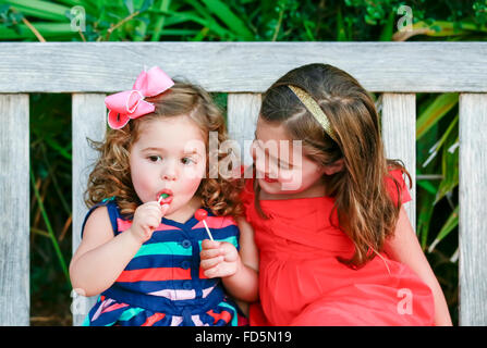 Two little girls in brightly colored dresses sitting on a bench enjoying lollipops. Stock Photo