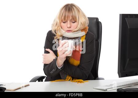 Cold businesswoman drinking a mug of hot coffee as she sits shivering at her desk in a knitted winter scarf Stock Photo