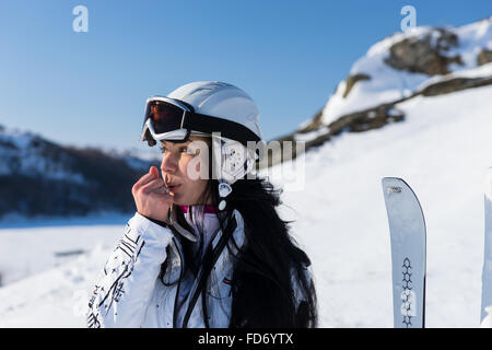 Close Up Head and Shoulders of Woman Wearing Helmet and Taking a Break from Skiing to Blow Hot Breath on Hands to Warm Them Up on Snow Covered Mountainside. Stock Photo