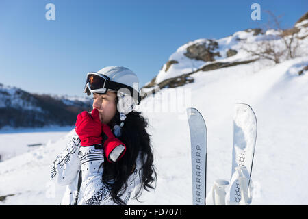 Waist Up of Young Woman with Long Dark Hair Wearing Ski Goggles and Helmet Standing Next to Skis and Warming Hands with Breath While Looking into the Distance on Sunny Snowy Mountainside. Stock Photo