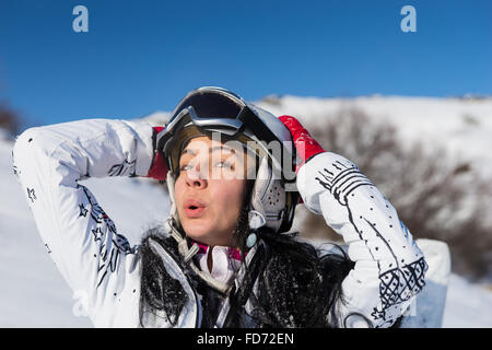 Head and Shoulders Close Up Playful Young Woman Wearing Helmet and Ski Goggles Standing on Snowy Mountainside with Hands Behind Head Enjoying Warm Sunshine on Beautiful Day. Stock Photo