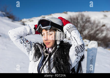 Waist Up Portrait of Young Woman with Long Dark Hair Standing with Skis and Wearing Helmet and Goggles with Hands Behind Head on Sunny Day on Snow Covered Mountainside. Stock Photo
