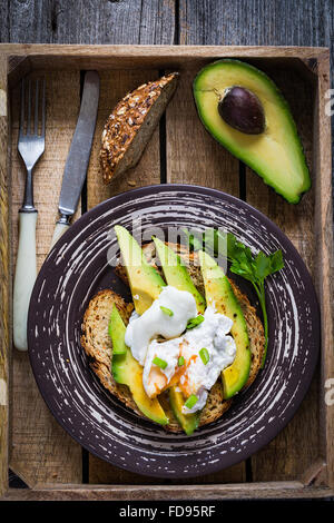 Poached egg and avocado on toasted whole wheat bread with seeds on a plate in vintage wooden tray. Garnished with scallions Stock Photo