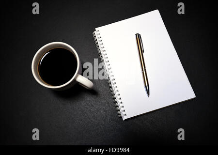 Blank notepad with office supplies and cup of coffee on black table. Above view and focus on the notebook Stock Photo