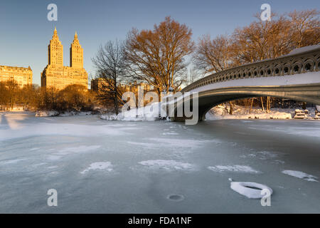 Cold Central Park winter sunrise on the frozen Lake with the Bow Bridge and Upper West Side buildings. Manhattan, New York City Stock Photo