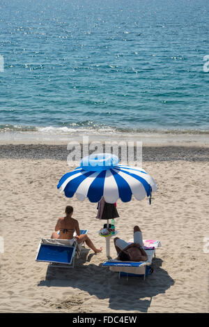 Young couple on sun loungers with blue and white parasol and swim ring on top on the Mingardo beach,Cilento,Campania,Italy Stock Photo
