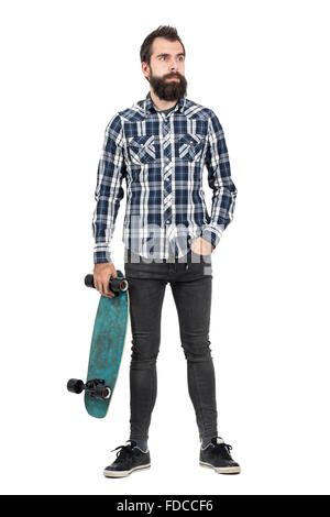 Confident hipster carrying skateboard in one hand looking away. Full body length portrait isolated over white studio background Stock Photo