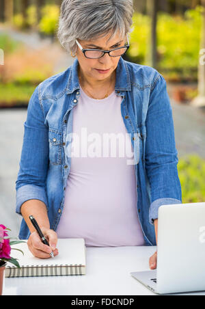Beautiful mature woman working in a greenhouse holding flowers and taking notes Stock Photo