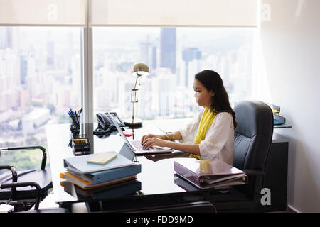 Adult businesswoman sitting in modern office in skyscraper. The secretary writes on laptop pc with serious expression. Wide shot Stock Photo