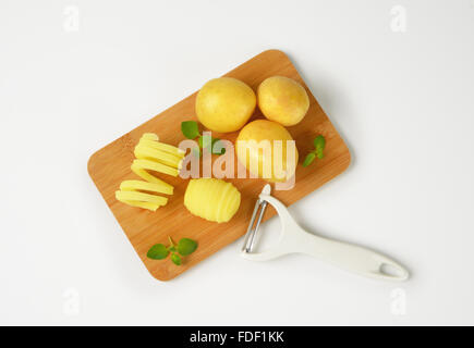 raw potatoes and peeler on wooden cutting board Stock Photo