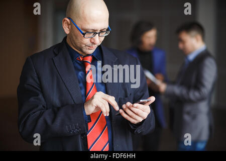 Portrait of male with glasses bald businessman using phone in hand, dressed striped suit and red tie, stands indoors office building. At background man discussing. Stock Photo
