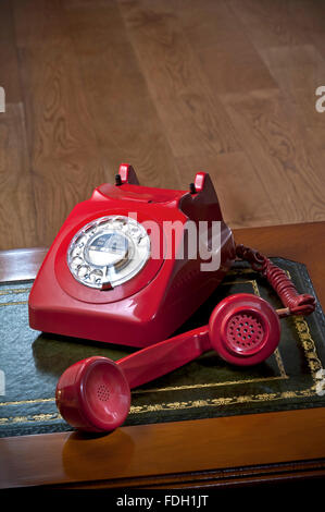 Classic retro red telephone with receiver 'off the hook' on desk with wooden floors behind Stock Photo