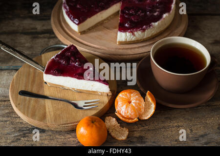Slice of cheesecake with cherries on wooden board, some mandarins and cup of tea Stock Photo