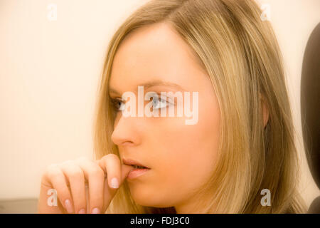 Teenage Caucasian girl with long blonde hair and blue eyes, looking at (unseen) item, with anxious expression, and nervously biting thumb. Stock Photo