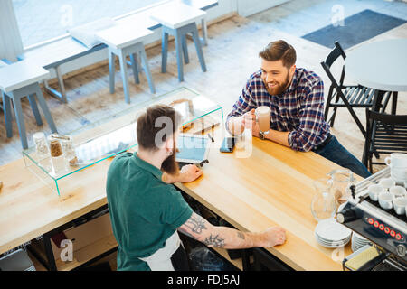 Cheeful attractive young man drinking coffee in cafeteria and talking to barista Stock Photo