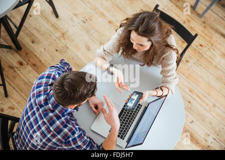 Top view of young happy couple sitting at round table in cafe and looking at screen of mobile phone and lapop Stock Photo