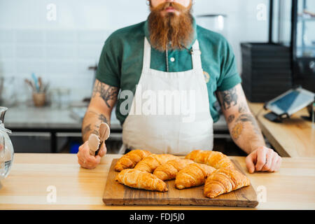 Closeup of bearded man in white apron with tattoo on hands standing in cafe and holding tongs for croissants Stock Photo
