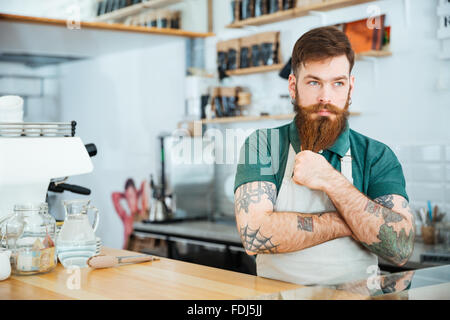 Handsome pensive man barista touching his beard and thinking in coffee shop Stock Photo