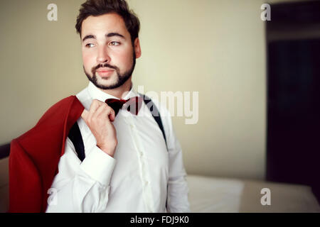 Stylish groom in tuxedo suit marsala red, burgundy bow tie, professional hairstyle, beard, mustache. Wedding preparations, getting ready. Man holding jacket over shoulder. Copy space for text. Stock Photo