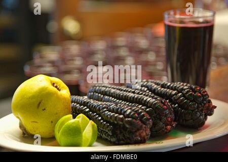 Peru, Lima, three purple maize corn cobs, a quince, and a sliced lime on a plate plate, glass of chicha morada in the background Stock Photo