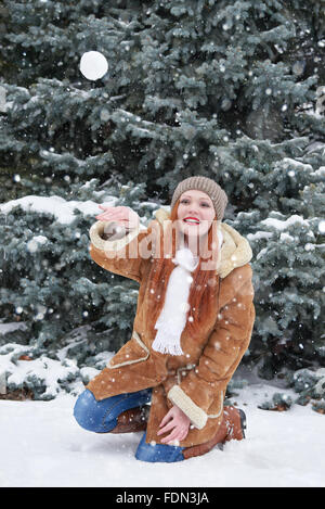 Girl play snowballs in winter forest at day. Snowy fir trees. Redhead woman full length. Stock Photo