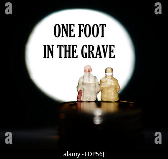 Miniature figurine pensioners sitting on coins in front of a spotlight showing the words One foot in the grave Stock Photo