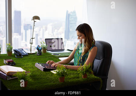 Concept of ecology and environment: Young business woman working in modern office with table covered of grass and plants. She ty Stock Photo